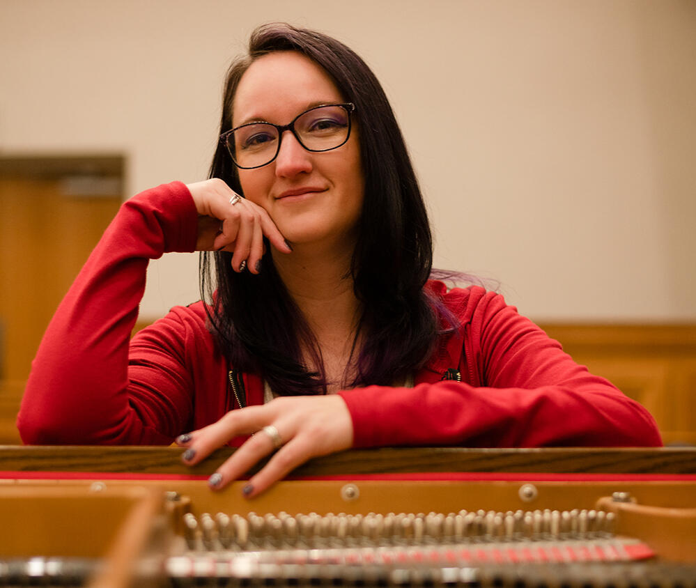 A woman with long brown hair and glasses sits confidently at a piano, with her head resting on her hand.