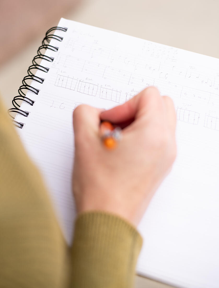 The focus is on a spiral notebook with staff lines on it, and my hand is shown writing musical notes in pencil.