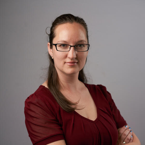 A woman with long brown hair and glasses, smiles confidently into the camera.